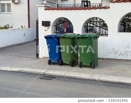 Waste containers lined up on a sidewalk near a residential wall. Recycling, sanitation, garbage collection, household disposal, urban maintenance, and neighborhood cleanliness. Waste containers lined up on a sidewalk near a residential wall. Recycling, sanitation, garbage collection, household disposal, urban maintenance, and neighborhood cleanliness. 139191445