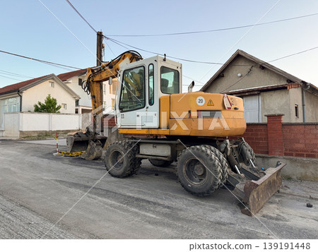 Wheeled excavator parked beside houses during road reconstruction work. Civil engineering, street maintenance, asphalt repair, utility access, heavy equipment, and residential infrastructure. Wheeled excavator parked beside houses during road reconstruction work. Civil engineering, street maintenance, asphalt repair, utility access, heavy equipment, and residential infrastructure. 139191448