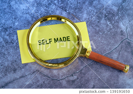 A golden magnifying glass with a wooden handle rests on a yellow sticky note, clearly showing the words SELF MADE on a dark gray cracked surface A golden magnifying glass with a wooden handle rests on a yellow sticky note, clearly showing the words SELF MADE on a dark gray cracked surface 139191650
