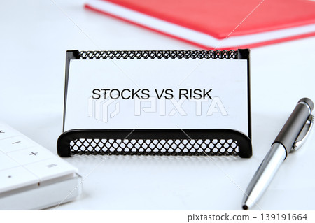 Desk: STOCKS VS RISK card, calculator, pen. Blurred red notebook behind. Signifies investment considerations Desk: STOCKS VS RISK card, calculator, pen. Blurred red notebook behind. Signifies investment considerations 139191664