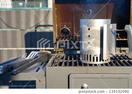 Charcoal chimney starter smokes on grill grate with tongs and brush nearby 139192090
