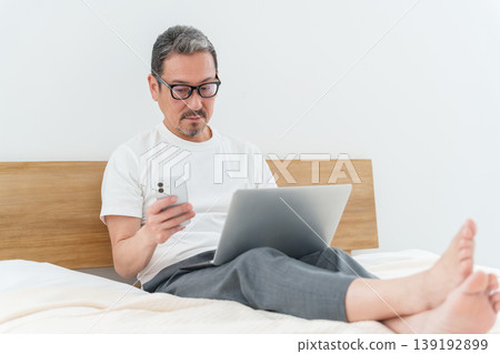 中年男子在床上使用智慧型手機和電腦。 中年男子在床上使用智慧型手機和電腦。 139192899