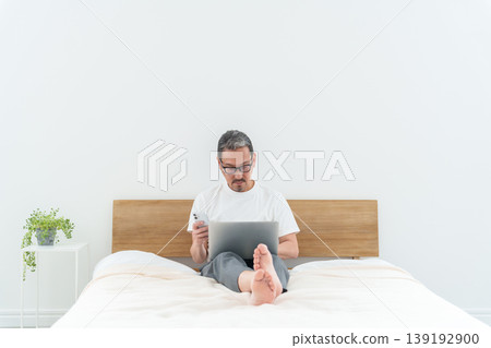 中年男子在床上使用智慧型手機和電腦。 139192900