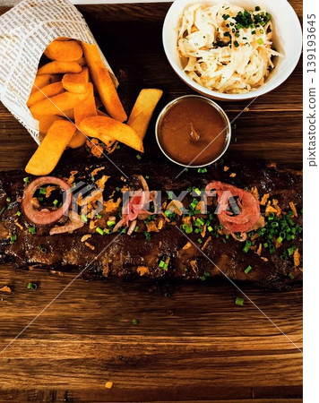 Grilled ribs garnished with diced green onions and red onion slices rest on a wooden platter. Potato fries, barbecue sauce, coleslaw Grilled ribs garnished with diced green onions and red onion slices rest on a wooden platter. Potato fries, barbecue sauce, coleslaw 139193645