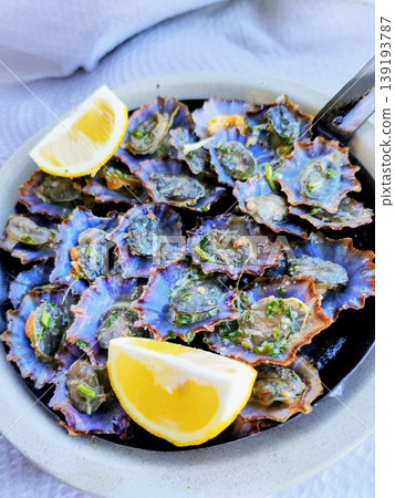 A plate filled with limpets (Patella spp.) is garnished with fresh lemon wedges. The barnacles are cooked, with herbs and spices on their surfaces.  139193787