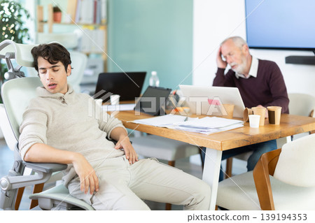 Tired elderly and young male colleagues fall asleep while working Tired elderly and young male colleagues fall asleep while working 139194353