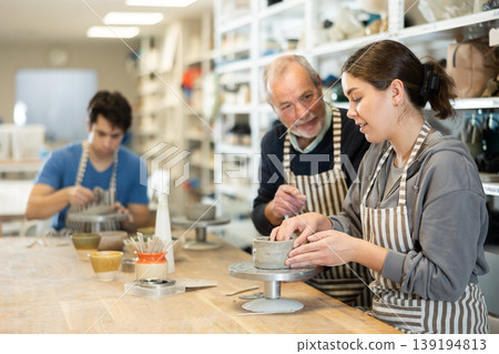 Elderly man teacher shows girl how to sculpt ceramic product from raw clay in workshop 139194813
