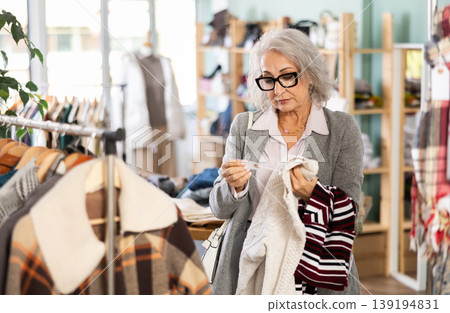 Mature female buyer in glasses chooses fashionable sweater in clothing store Mature female buyer in glasses chooses fashionable sweater in clothing store 139194831