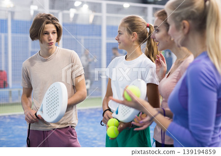Teenage boy, girl and adult women standing in padel court and talking Teenage boy, girl and adult women standing in padel court and talking 139194859