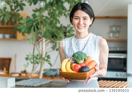 一位身穿運動服的年輕亞洲女性在廚房前拿著蔬菜（缺乏蔬菜、營養平衡、綠色和黃色蔬菜） 139195033