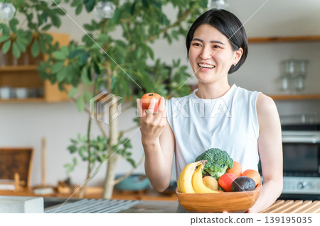 一位身穿運動服的年輕亞洲女性在廚房前拿著蔬菜（缺乏蔬菜、營養平衡、綠色和黃色蔬菜） 139195035