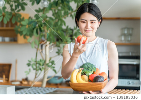 一位身穿運動服的年輕亞洲女性在廚房前拿著蔬菜（缺乏蔬菜、營養平衡、綠色和黃色蔬菜） 139195036