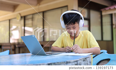 Asian Boy Student Wearing Headphones Studying Online with Laptop and Writing on Notebook Asian Boy Student Wearing Headphones Studying Online with Laptop and Writing on Notebook 139197173