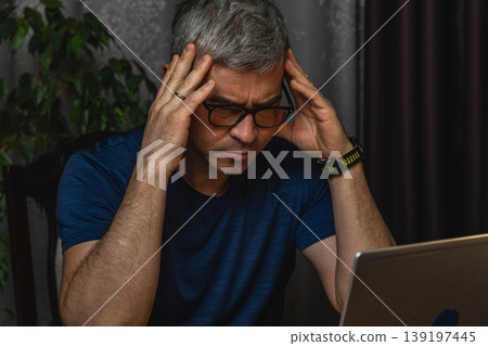 Overworked man holding head while working on laptop at night Overworked man holding head while working on laptop at night 139197445