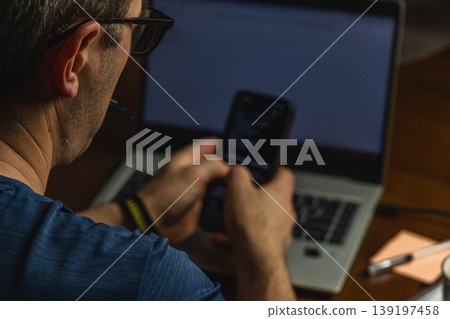 Man using smartphone while working on laptop from home Man using smartphone while working on laptop from home 139197458