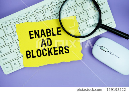 Yellow torn paper with the text "Enable Ad Blockers" placed on a white keyboard, and magnifying glass and a computer mouse 139198222
