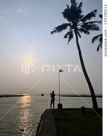 A silhouette of a person photographing a backwater with palm trees and a sunset in the background at a resort in Kerala, South India. A silhouette of a person photographing a backwater with palm trees and a sunset in the background at a resort in Kerala, South India. 139199213