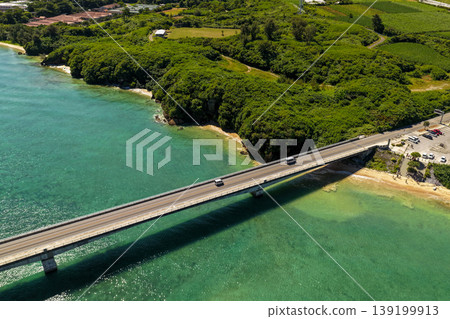 An aerial photograph taken in midsummer showing Kouri Bridge, the emerald green sea, and lush green cliffs and a small beach. An aerial photograph taken in midsummer showing Kouri Bridge, the emerald green sea, and lush green cliffs and a small beach. 139199913