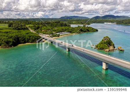 A photograph taken from the midsummer sky, showing Kouri Bridge and a small island floating in the emerald green sea. A photograph taken from the midsummer sky, showing Kouri Bridge and a small island floating in the emerald green sea. 139199946