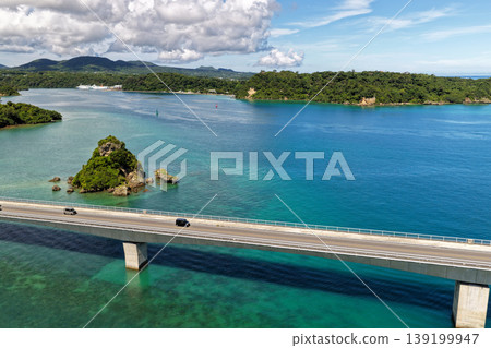 A photograph taken from the midsummer sky, showing Kouri Bridge and a small island floating in the emerald green sea. A photograph taken from the midsummer sky, showing Kouri Bridge and a small island floating in the emerald green sea. 139199947