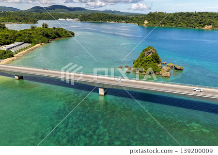 A photograph taken from the midsummer sky, showing Kouri Bridge and a small island floating in the emerald green sea. A photograph taken from the midsummer sky, showing Kouri Bridge and a small island floating in the emerald green sea. 139200009