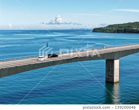 A summer day photograph, framed horizontally, showing a car driving across the emerald blue sea and the Kouri Bridge. 139200046
