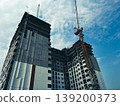 Low angle view of a high-rise building under construction with tower crane reaching into blue sky, modern concrete structure rising, urban development concept, architecture progress and industrial 139200373