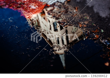 A striking reflection of a unique, old urban building in a puddle after rain Moody high-contrast image captures architectural details and the sky in the water's surface, highlighted by a splash of red A striking reflection of a unique, old urban building in a puddle after rain Moody high-contrast image captures architectural details and the sky in the water's surface, highlighted by a splash of red 139200412