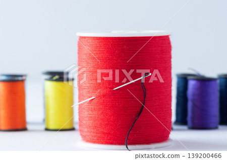 A spool of red thread with a needle, set against a background of spools of colored thread. 139200466