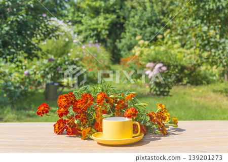 Summer still life; yellow cup of tea and bouquet of marigold flowers on a wooden table in a blooming summer garden by sunny day Summer still life; yellow cup of tea and bouquet of marigold flowers on a wooden table in a blooming summer garden by sunny day 139201273