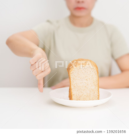 Unhappy woman showing thumbs down gesture and refusing to eat bread. concept of gluten intolerance, gluten sensitivity, carb loading concerns, or a disliking for unhealthy dietary habits Unhappy woman showing thumbs down gesture and refusing to eat bread. concept of gluten intolerance, gluten sensitivity, carb loading concerns, or a disliking for unhealthy dietary habits 139201656
