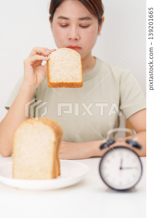Happy smiling woman eating whole wheat bread with alarm clock. concept of intermittent fasting, IF, meal timing, and choosing healthy carbohydrates for a balanced diet and weight loss management Happy smiling woman eating whole wheat bread with alarm clock. concept of intermittent fasting, IF, meal timing, and choosing healthy carbohydrates for a balanced diet and weight loss management 139201665