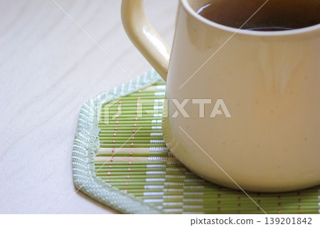 Close up of a warm ceramic mug filled with tea on a green bamboo coaster on a light wooden surface 139201842
