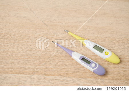 Two colorful digital thermometers rest on a light wooden surface, suggesting fever detection, home health care and illness monitoring. Clear LCD displays practical everyday temperature measurement. Two colorful digital thermometers rest on a light wooden surface, suggesting fever detection, home health care and illness monitoring. Clear LCD displays practical everyday temperature measurement. 139201933