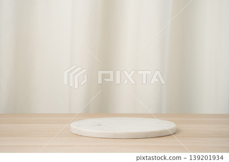 Minimal white marble pedestal placed on a light wooden table with a soft neutral curtain backdrop, ideal for product display, studio mockups, presentations, and clean commercial staging. 139201934