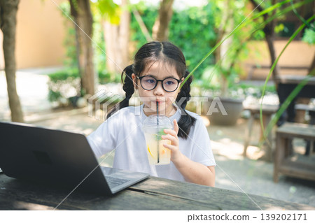 Young girl with glasses sitting at a wooden table using laptop while enjoying a refreshing drink in a sunny outdoor garden setting Young girl with glasses sitting at a wooden table using laptop while enjoying a refreshing drink in a sunny outdoor garden setting 139202171