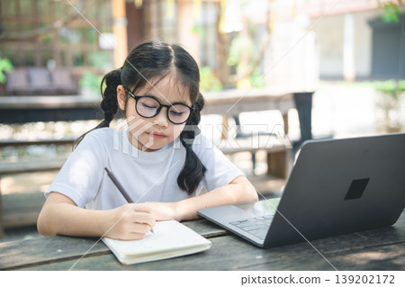 Young girl with glasses writes notes in a notebook while learning at an outdoor table near a laptop in a sunny garden setting Young girl with glasses writes notes in a notebook while learning at an outdoor table near a laptop in a sunny garden setting 139202172