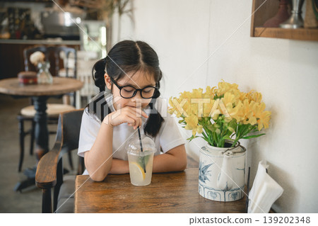 Young girl with glasses enjoying refreshing lemon drink in cozy cafe with beautiful yellow flowers on wooden table, perfect for lifestyle and family themes Young girl with glasses enjoying refreshing lemon drink in cozy cafe with beautiful yellow flowers on wooden table, perfect for lifestyle and family themes 139202348