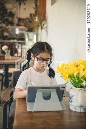 Young girl with glasses enjoying refreshing lemon drink in cozy cafe with beautiful yellow flowers on wooden table, perfect for lifestyle and family themes 139202349