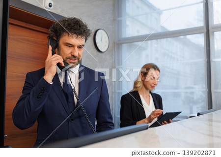 Male receptionist talking on phone while colleague works with tablet at hotel desk 139202871
