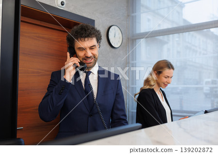 Smiling hotel front desk manager speaking on telephone in modern lobby 139202872