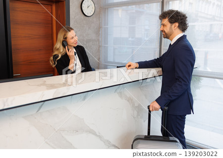 Professional Receptionist Speaking on Phone While Welcoming Business Guest at Hotel Front Desk Professional Receptionist Speaking on Phone While Welcoming Business Guest at Hotel Front Desk 139203022