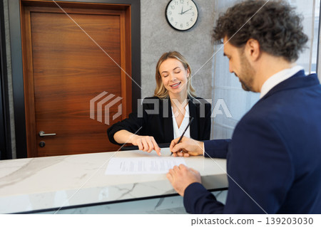 Receptionist Pointing at Contract While Guest Signs Paperwork at Modern Hotel Lobby Desk Receptionist Pointing at Contract While Guest Signs Paperwork at Modern Hotel Lobby Desk 139203030