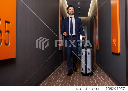 Businessman walking in hotel corridor with suitcase toward room door Businessman walking in hotel corridor with suitcase toward room door 139203057