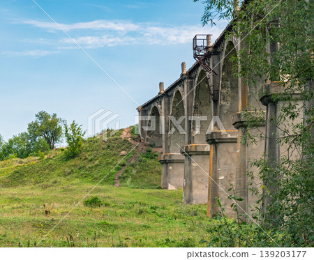Tall weathered concrete viaduct arches majestically over a vibrant green field on a clear day Tall weathered concrete viaduct arches majestically over a vibrant green field on a clear day 139203177