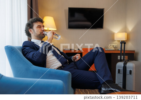 Tired businessman drinking water while resting in hotel room after long business trip Tired businessman drinking water while resting in hotel room after long business trip 139203711