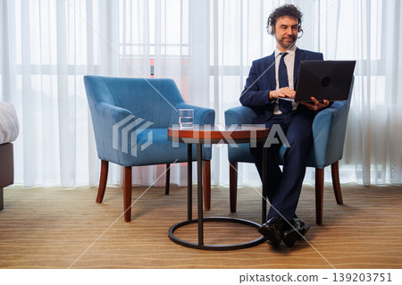Remote Worker in Business Suit Using Laptop with Headset in Hotel Room Remote Worker in Business Suit Using Laptop with Headset in Hotel Room 139203751