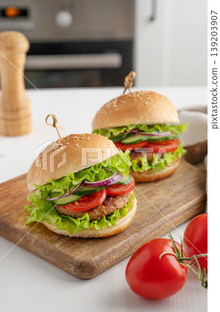 Two beef burgers with tomato, cheese, and lettuce on a cutting board on a light table Two beef burgers with tomato, cheese, and lettuce on a cutting board on a light table 139203907