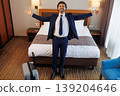 Excited businessman standing in hotel room with open arms in front of bed 139204646