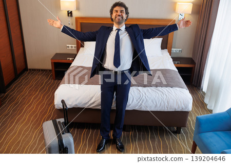 Excited businessman standing in hotel room with open arms in front of bed Excited businessman standing in hotel room with open arms in front of bed 139204646